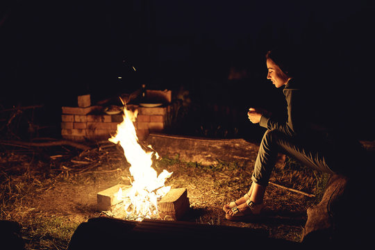 Young woman in geans and hoodie in the mountains. Young woman with fire on mountain at sunset. Girl standing near a fireplace, back view
