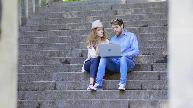 Couple with laptop sitting on stairs in town talking.
