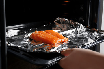Female hand putting baking sheet with salmon on foil into oven
