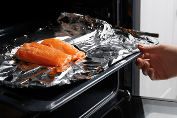 Female hand putting baking sheet with salmon on foil into oven