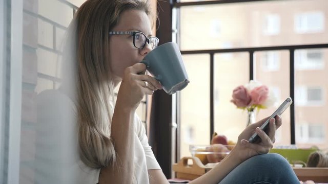 A young woman is sitting on a cozy terrace, holding a mug with coffee and a smartphone. Morning coffee on the balcony: a pretty girl uses a smartphone against the a brick wall and looks out street