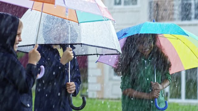PAN of laughing little boys and African-American girl with curly hair wearing raincoats and holding umbrellas while standing under pouring rain