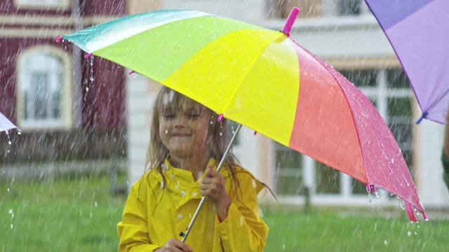 PAN of cute little girl in yellow raincoat holding umbrella and laughing while jumping in rain with unrecognizable friends