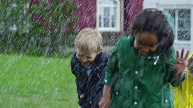 Slow motion of happy little children with umbrellas wearing raincoats running on green grass in heavy rain