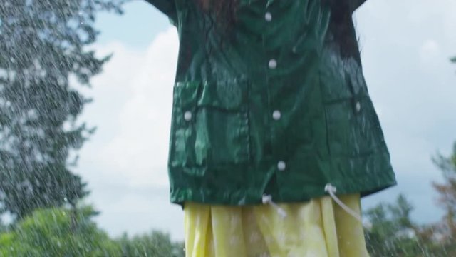 Tilt down of excited little African American girl in green rubber boots and raincoat jumping in puddle in rain