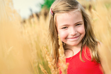 Portrait a happy little smile girl in red dress among high field grass at summer day