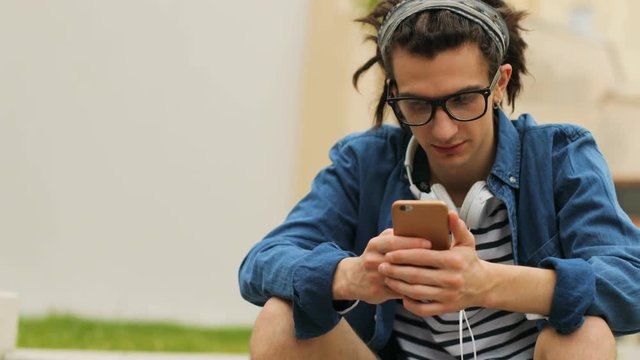 Stylish hipster man with dreadlocks using smart phone for surfing in the internet during resting in the beatiful city.