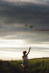 Back view of girl holding kite and running in field under sky with overcast.