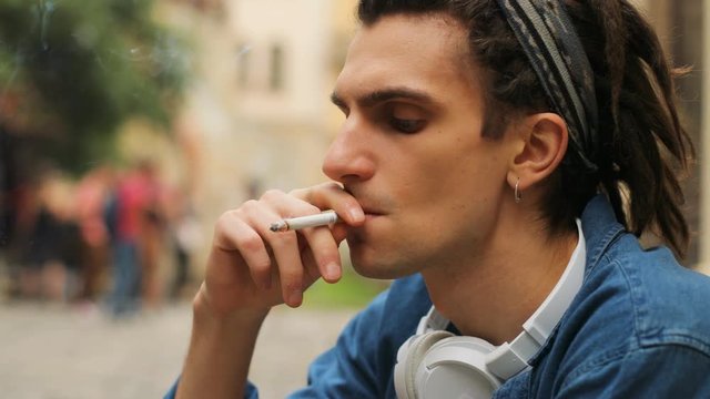 Portrait of hipster man with dreadlocks smoking cigarette while sitting on the bench in the city centre. Side view.