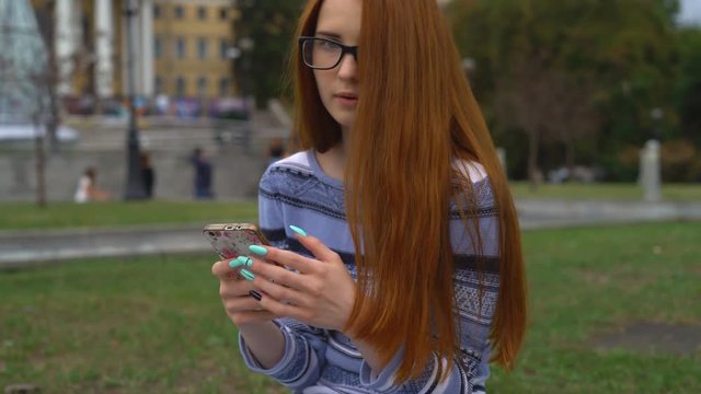 Young attractive teenage girl on a walk in the city. The woman has long red hair and glasses. The girl touches her long hair and smiles. A student sits in social networks through a mobile phone.