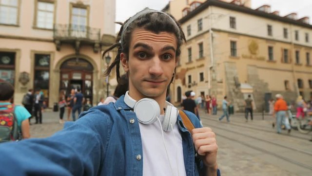 Young hipster man making funny faces on the camera while he making selfie on the street background. Portrait shot.