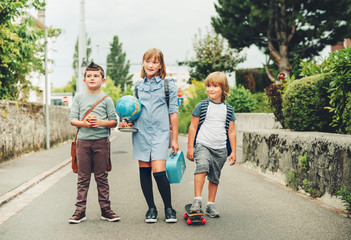 Group of three funny kids wearing backpacks walking back to school. Girl and boys enjoying school activities. Globe, lunch box, red apple and skateboard accessories.