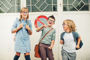 Group of three funny kids wearing backpacks walking back to school. Girl and boys wearing eyeglasses posing outdoors