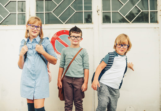 Group Of Three Funny Kids Wearing Backpacks Walking Back To School. Girl And Boys Wearing Eyeglasses Posing Outdoors