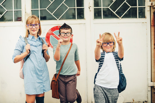 Group Of Three Funny Kids Wearing Backpacks Walking Back To School. Girl And Boys Wearing Eyeglasses Posing Outdoors