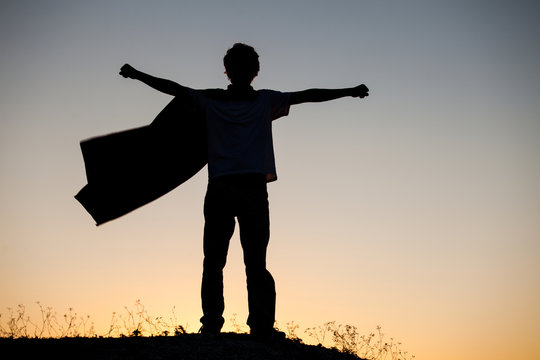 Boy playing superheroes on the sky background, silhouette of teen superhero in a raincoat on the hill