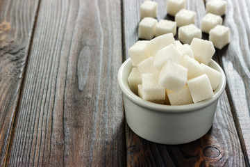 White sugar in bowl on wooden background. Selective focus, horizontal. A few sugar cubes are near the full glass with white sugar. Intake of bad calories.
