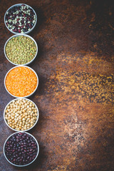 Bowls of various lagumes - black, white, red beans, red, green lentils, and chickpeas on old rusty background. Top view, space for text, toned.
