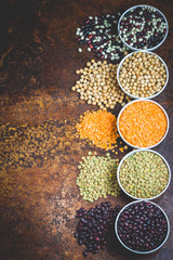Various lagumes in a bowls - black, white, red beans, red, green lentils, and chickpeas on dark stone  background. Top view, space for text, toned.