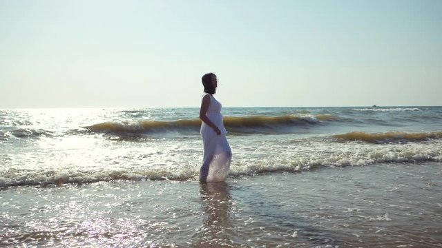 Gypsy young brunette girl in white maxi long dress walking on the waves in sea or ocean. Windy beach. Bohemian lifestyle. Slow motion. Film tonned