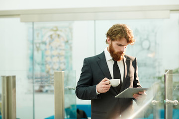 Bearded trader with cup of tea and tablet reading online news in the morning