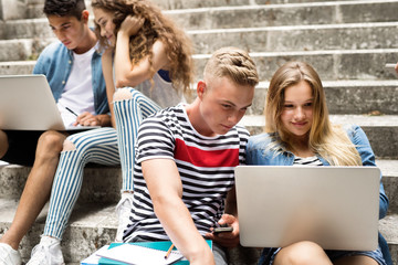 Teenage students with laptops sitting on stone steps.