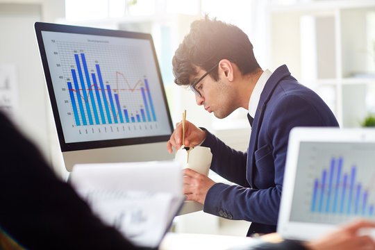 Busy handsome manager in suit eating instant noodles with chopsticks while sitting at desk in open plan office, profile view