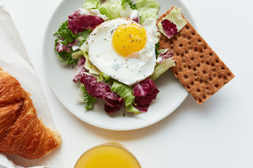 Served table for breakfast with freshly baked croissant, glass of orange juice and plate with lettuce, fried egg and homemade crispbread, directly above view