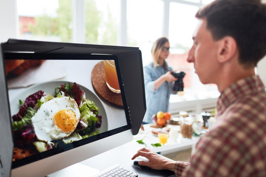 Profile view of concentrated young food designer sitting in front of computer monitor and retouching photos, his female colleague taking pictures of appetizing breakfast