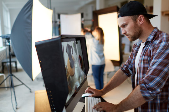 Profile view of young handsome food designer retouching photo while standing in front of modern computer, interior of production studio on background