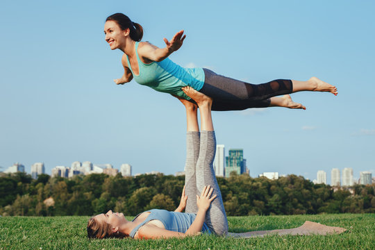 Two young beautiful Caucasian women yogi doing acro bird yoga pose. Women doing stretching workout in park outdoors at sunset. Healthy lifestyle modern activity