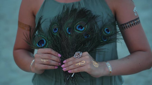 Hands of lady at beach with flash tattoos holding and touching peacock feathers fan. Gypsy boho style. Slow motion. Beautiful scene, film tonned.