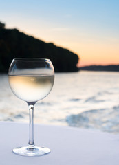 glass of cool white wine on white tablecloth with coastline in blurred background at sunset