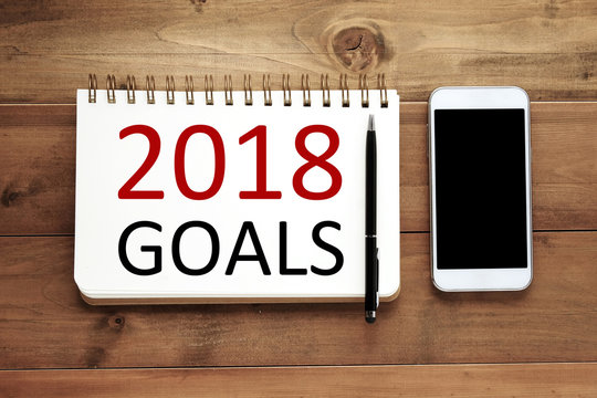 2018 goals on paper note book background and smart phone on wood table, business and new year concept