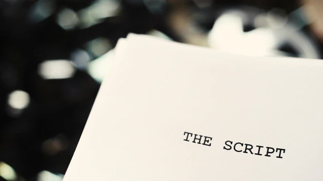 White cover paper with script title text close up over blurred unrolled movie filmstrip background