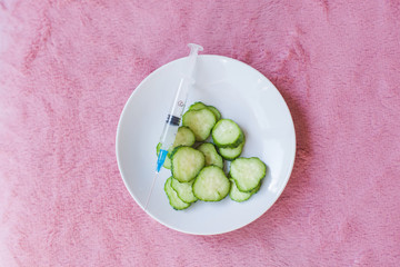 White plate with slices of green cucumber and a syringe with an injection. Top view. Pink background.