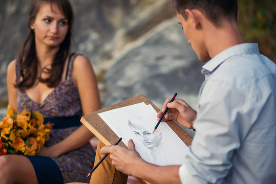 Young lovely couple on a romantic open-air date. A young man paints a portrait of his beloved girl sitting on a rock. Graphic portrait close up.
