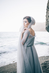 Fototapeta na wymiar Beautiful girl bride in a long grey dress with lace,with a veil on the beach, closed with a veil, viewing profile