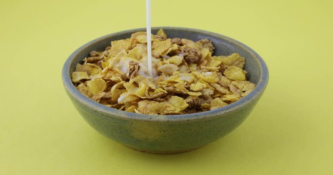 A full bowl with multigrain breakfast cereal with skim milk being poured onto the food with one splatter atop a yellow tabletop..