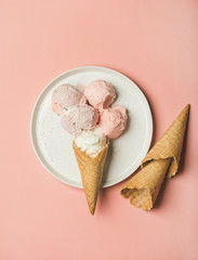 Flatlay of pastel pink strawberry and coconut ice cream scoops and sweet cones on white plate over pastel pink background, top view, copy space, vertical composition