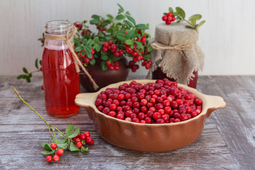 Very ripe cowberries in a bowl on the table and cranberry drinks and jam. The bouquet of sprigs of cranberries in a pot. On a wooden background, space for text.