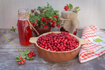Very ripe cowberries in a bowl on the table and cranberry drinks and jam. The bouquet of sprigs of cranberries in a pot. On a wooden background, space for text.