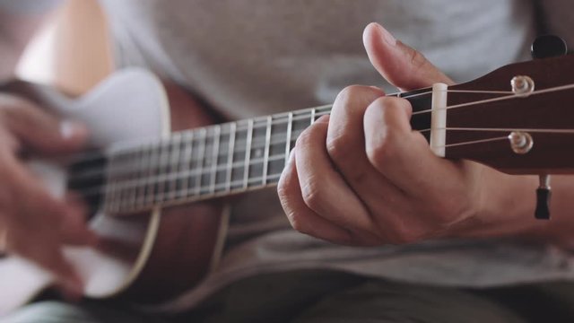 Slow dolly crop shot of man holding and playing acoustic ukulele guitar sitting inside.