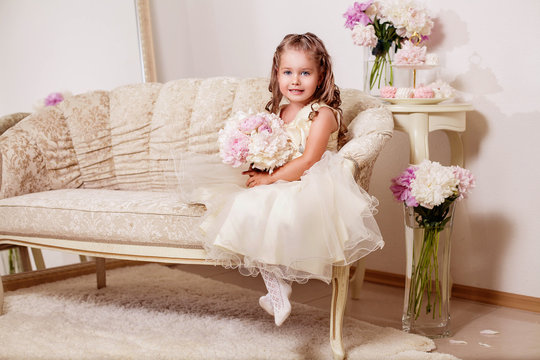 Happy smiling flower girl sitting on the beige sofa with bouquet of peony flowers and sweets. Wedding. Just married.