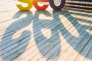 The word SEO by large colored letters creating a shadow on the wooden terrace on the river bank. Sun backlight. Selective focus. Space for text
