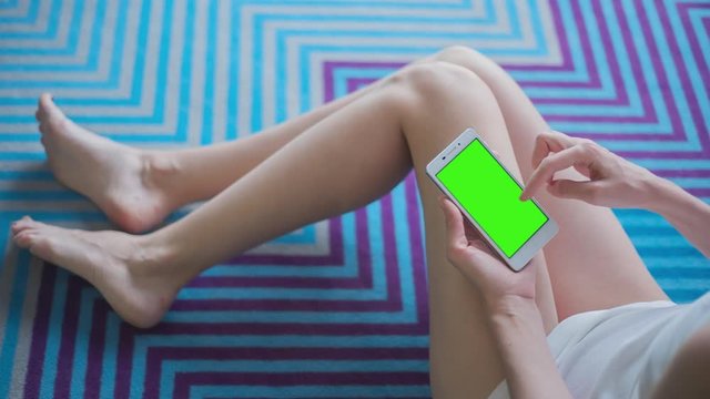 Young Woman in white top sitting on the pattern floor uses mobile phone with pre-keyed green screen. Few types of gestures - scrolling up and down, tapping, zoom in and out