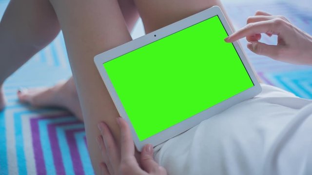 Young Woman in white top sitting on the pattern floor uses Tablet PC with pre-keyed green screen. Few types of gestures - scrolling up and down, tapping, zoom in and out