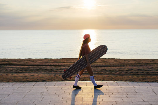 Teen girl holding her longboard by beach at sunrise