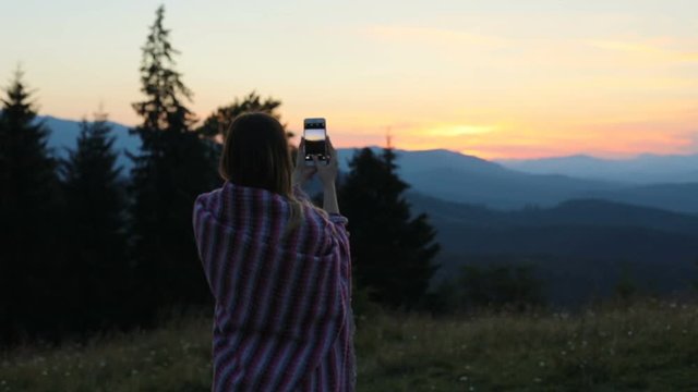Woman girl holding smart phone taking photo picture standing back view sunset background fiery colored sky photograph landscape rest enjoying nature vacation summer evening memory wrapped blanket dusk