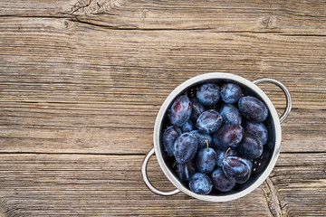 Fresh blue plums in rustic colander on old wooden table. Organic food. Top view, copy space.